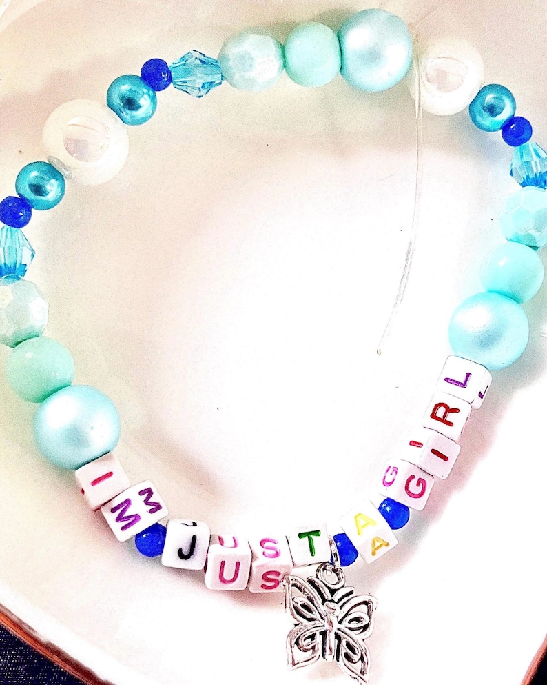 May include: A beaded bracelet featuring a mix of light blue, turquoise, and white round beads, interspersed with small dark blue beads. White square letter beads spell out the phrase I AM JUST A GIRL, accented by a silver-toned butterfly charm.