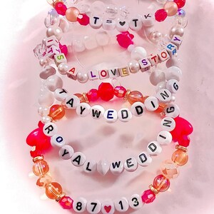 May include: A stack of four elastic beaded bracelets featuring white letter beads and colorful accent beads. The bracelets display the text TS heart T, IT'S A LOVE STORY, TAYWEDDING, ROYAL WEDDING, and 87 heart 13, interspersed with pink, orange, and clear beads.