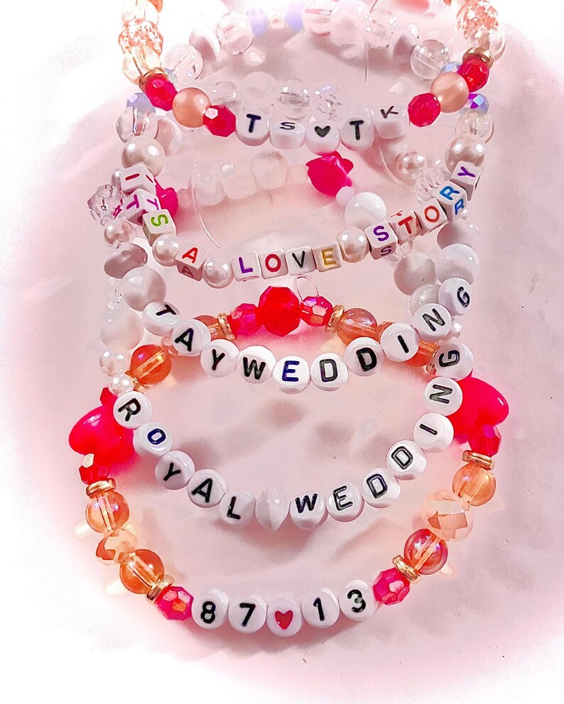 May include: A stack of four elastic beaded bracelets featuring white letter beads and colorful accent beads. The bracelets display the text TS heart T, IT'S A LOVE STORY, TAYWEDDING, ROYAL WEDDING, and 87 heart 13, interspersed with pink, orange, and clear beads.