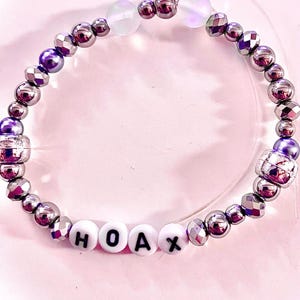 K&ouml;nnte beinhalten: Ein Perlenarmband mit metallisch silbernen und violetten facettierten Perlen, zwei durchscheinenden irisierenden Perlen und vier wei&szlig;en runden Perlen mit den schwarzen Buchstaben H, O, A und X, aufgereiht auf einem transparenten Gummiband.