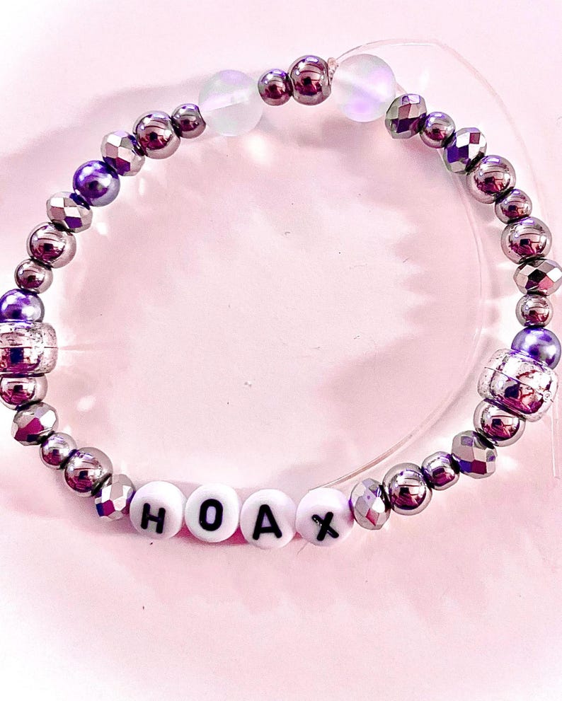 K&ouml;nnte beinhalten: Ein Perlenarmband mit metallisch silbernen und violetten facettierten Perlen, zwei durchscheinenden irisierenden Perlen und vier wei&szlig;en runden Perlen mit den schwarzen Buchstaben H, O, A und X, aufgereiht auf einem transparenten Gummiband.