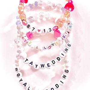 May include: A stack of four beaded bracelets featuring white letter beads spelling out 87 13, IT'S A LOVE STORY, TAYWEDDING, and ROYAL WEDDING. The bracelets include clear, iridescent, and pink heart-shaped beads, along with white pearl-like beads.