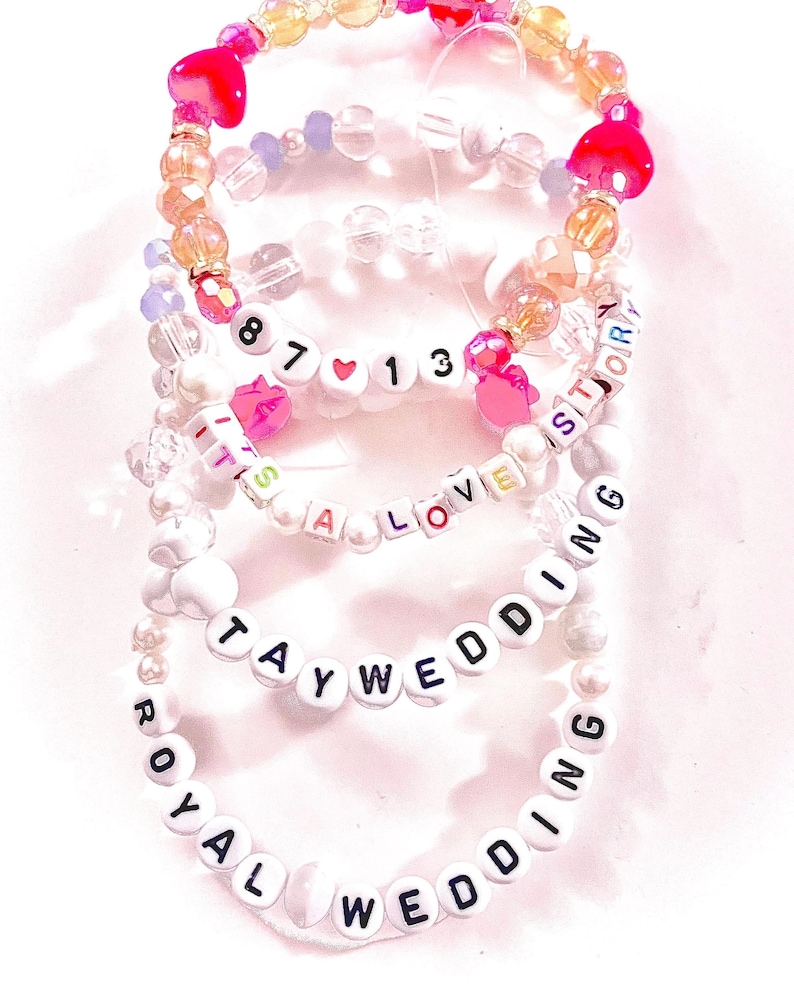 May include: A stack of four beaded bracelets featuring white letter beads spelling out 87 13, IT'S A LOVE STORY, TAYWEDDING, and ROYAL WEDDING. The bracelets include clear, iridescent, and pink heart-shaped beads, along with white pearl-like beads.