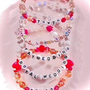 May include: A stack of five beaded bracelets featuring colorful plastic and clear beads. The bracelets include letter beads spelling out TS heart TK, IT'S A LOVE STORY, TAY WEDDING, ROYAL WEDDING, and 87 heart 13. The beads are in shades of pink, white, and orange.