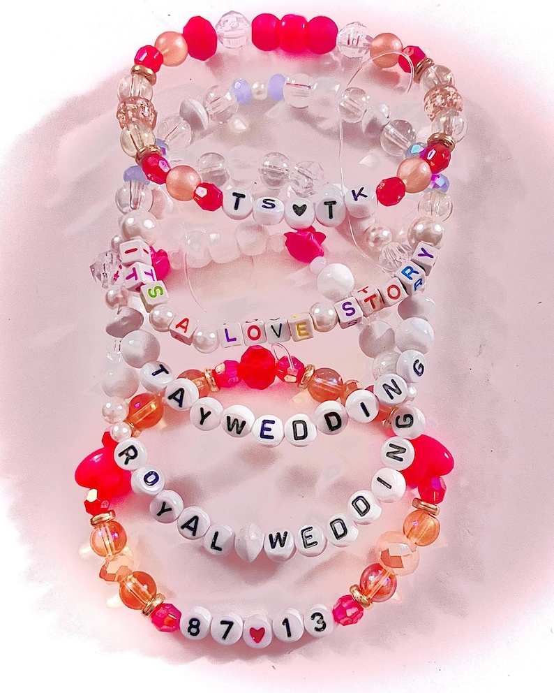 May include: A stack of five beaded bracelets featuring colorful plastic and clear beads. The bracelets include letter beads spelling out TS heart TK, IT'S A LOVE STORY, TAY WEDDING, ROYAL WEDDING, and 87 heart 13. The beads are in shades of pink, white, and orange.