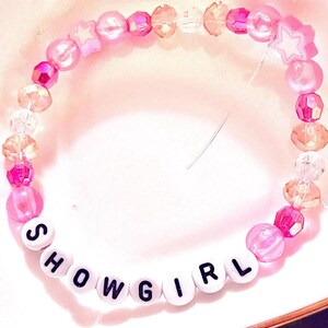 A handmade Taylor Swift The Life of a Showgirl inspired friendship bracelet featuring the word “Showgirl.” Designed with pink glass and plastic beads, this elastic bracelet sparkles with letter beads that capture the glamorous energy of the album’s title track. Perfect for Swifties to wear to concerts, stack with other bracelets, or gift to a fan. Trendy Taylor Swift bracelet inspired by The Life of a Showgirl era, ideal for collectors and concert accessories