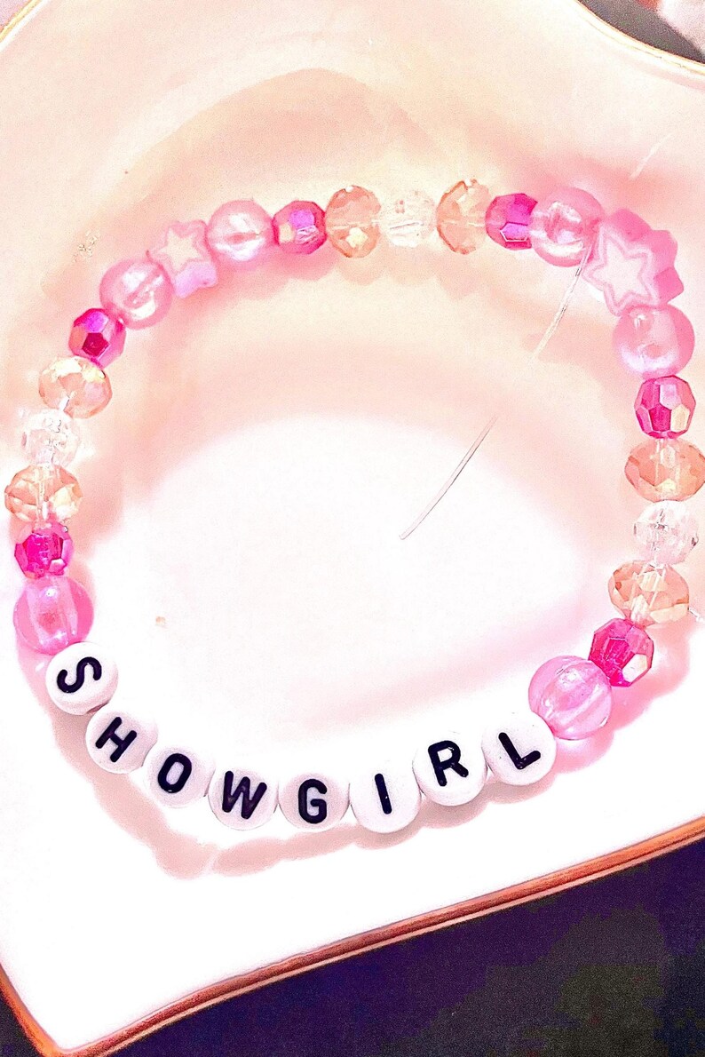 A handmade Taylor Swift The Life of a Showgirl inspired friendship bracelet featuring the word “Showgirl.” Designed with pink glass and plastic beads, this elastic bracelet sparkles with letter beads that capture the glamorous energy of the album’s title track. Perfect for Swifties to wear to concerts, stack with other bracelets, or gift to a fan. Trendy Taylor Swift bracelet inspired by The Life of a Showgirl era, ideal for collectors and concert accessories