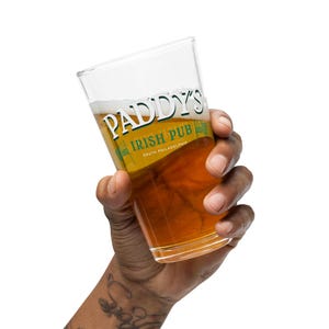 May include: A hand holding a glass of beer with a green and white label that reads "Paddy's Irish Pub South Philadelphia".