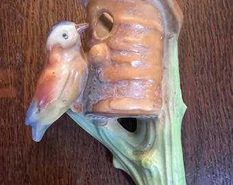 May include: A ceramic wall-mounted birdhouse figurine featuring a light brown cylindrical structure with a roof and a circular opening. A small bird with blue, white, and orange coloring is perched on the side, attached to a green tree branch base.