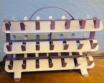 May include: A three-tiered, metal bottle drying rack with a handle. The rack is a muted rose gold color with dark brown bottle holders. The rack has a rectangular shape with a handle on top and is designed to hold multiple bottles.