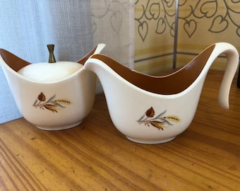 May include: A white ceramic creamer and sugar bowl set with a brown rim and a wheat and leaf design. The sugar bowl has a lid with a gold knob.