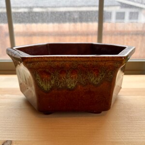 May include: A brown ceramic planter with a geometric shape. The planter has a glossy finish with a gradient of green and brown hues. It sits on small feet and is suitable for indoor or outdoor use.