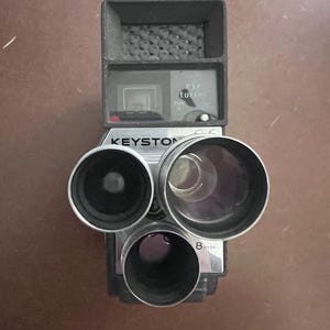 May include: Vintage Keystone 8mm movie camera. The camera is silver and black with three lenses and the text "8mm". The camera has a viewfinder and the word "Keystone" is printed on the front.