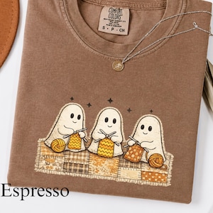 May include: A brown crewneck t-shirt featuring a graphic of three white ghosts knitting with yarn on a patchwork quilt. The shirt has a Comfort Colors brand label at the neck and a thin silver chain necklace resting on the fabric near the collar.