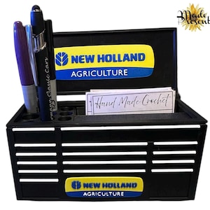 May include: Black desk tidy featuring the text "NEW HOLLAND AGRICULTURE" in yellow and blue. It has a pen holder and a slot for cards labelled "Hand Made Crochet". The "Made Present" logo is also visible.