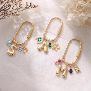 May include: Three gold-toned oval carabiner keychains featuring dangling charms. The first has a letter S, a blue shell, a blue stone, and a music note. The second has a letter A, a green clover, a green stone, and a flower. The third has a letter M, a pink glass, a red stone, and a bear.