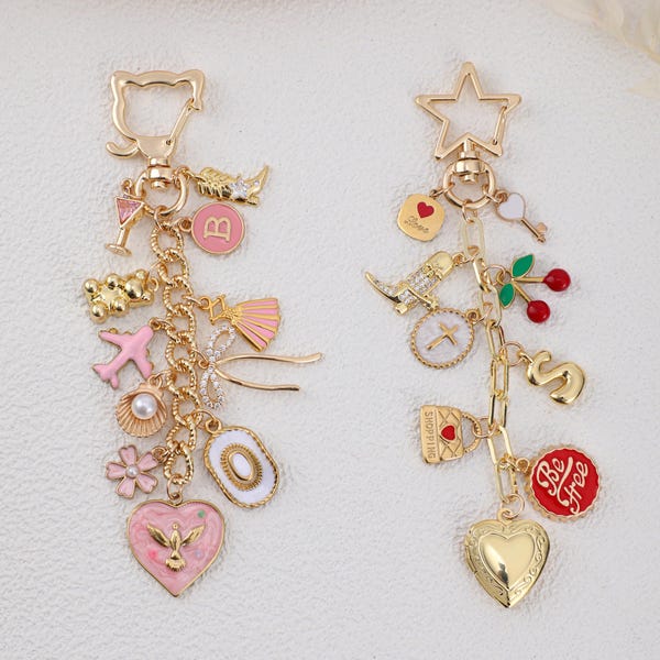 May include: Two gold-toned metal keychains featuring various charms. The left keychain has a cat-shaped clasp with charms including a pink heart with a bird, a shell, a plane, and a bear. The right keychain has a star-shaped clasp with charms including a heart locket, a red circle with the text 'Be Free', cherries, and a gold letter S.
