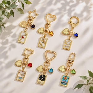 May include: Six gold-toned metal keychains featuring rectangular tarot card charms with enamel details, dangling heart-shaped charms with engraved letters, and additional coloured crystal heart accents. Each keychain has a unique clasp shape.