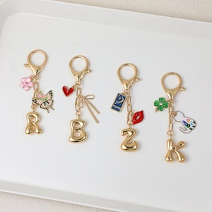 May include: Four gold-toned metal keychains featuring letters R, B, Z, and K. Each keychain has a lobster clasp and unique charms: a pink flower and butterfly for R, a red heart and bow for B, a blue moon card and red lips for Z, and a green clover and cat for K.