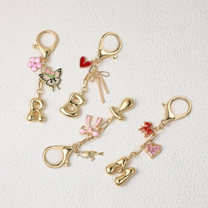 Pode incluir: Quatro porta-chaves de metal dourado com fechos de mosquetão. Um tem a letra R, uma flor rosa e uma borboleta. Um tem a letra B e um coração vermelho com um laço. Um tem a letra L e um laço rosa. Um tem a letra M, uma flor vermelha e um coração rosa.