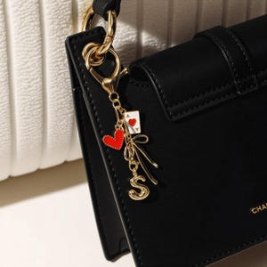 Puede incluir: Un charm dorado con un corazón rojo, una carta de juego de as de corazones y la letra "S" cuelga de un bolso negro. El charm está sujeto a la bolsa con un cierre dorado. El bolso es negro con un cierre dorado.