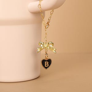 Pode incluir: Um colar dourado com um pingente de laço e um berloque em forma de coração preto. O berloque apresenta a letra "B" em dourado. O colar está pendurado em um fundo neutro.
