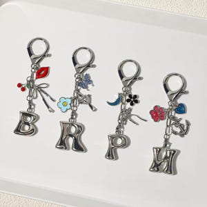 May include: Four silver-toned metal keychains featuring large, puffy letter charms B, R, P, and H. Each keychain includes a lobster clasp and various small enamel and metal charms, including a red lip, cherries, a blue flower, a butterfly, a moon, a black flower, a pink paw print, a blue heart, and an anchor.