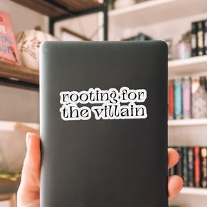 May include: A black e-reader with a white sticker that reads "rooting for the villain." The e-reader is held in front of a bookshelf filled with books. The sticker is in a serif font.