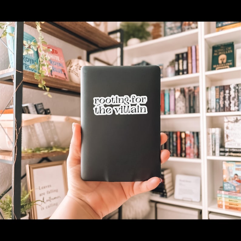 May include: A black e-reader with a white sticker that reads "rooting for the villain." The e-reader is held in front of a bookshelf filled with books. The sticker is in a serif font.