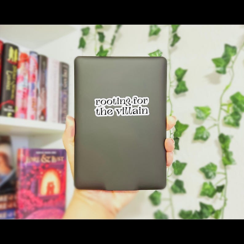 May include: A dark gray tablet with the text "rooting for the villain" on a white sticker. The tablet is held in a hand, with a bookshelf and green vines in the background.