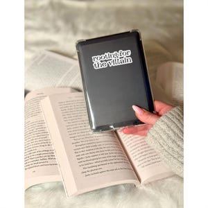 May include: A black e-reader case with the text "rooting for the villain" in white lettering. The e-reader is held over an open book with printed pages. The background is a soft, neutral color.