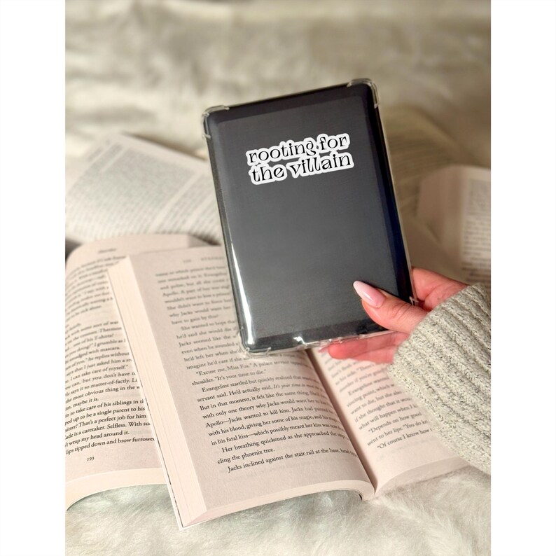 May include: A black e-reader case with the text "rooting for the villain" in white lettering. The e-reader is held over an open book with printed pages. The background is a soft, neutral color.