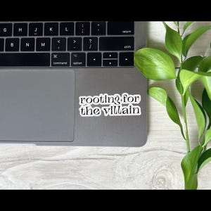 May include: A white sticker with the text "rooting for the villain" in a black, stylized font. The sticker is placed on a gray laptop, with a keyboard visible. Green leafy plant stems are in the background.