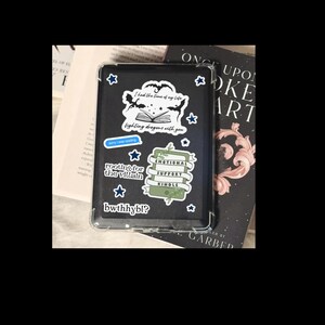 May include: A black Kindle e-reader with a clear protective case, decorated with various stickers. The stickers include phrases like "I had the time of my life," "fighting dragons with you," and "Emotional Support Kindle."