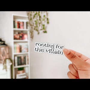 May include: A white sticker with the text "rooting for the villain" in a black, stylized font. The sticker is held by a hand against a white wall, with a bookshelf and hanging plant in the background.