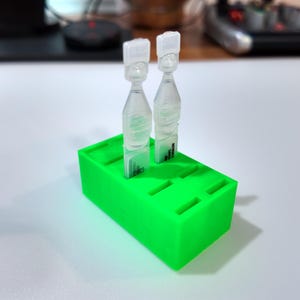 May include: Two clear plastic vials with white caps are displayed on a bright green rectangular holder. The holder has several slots for the vials. The background is a white surface with a blurred view of a desk and computer accessories.