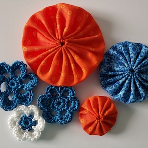 May include: Five fabric and crocheted flowers in shades of orange, blue, and white. The flowers are arranged on a white surface.