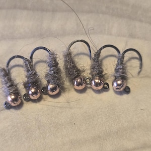 May include: Six fishing flies with gray bodies, black hooks, and rose gold-colored beads. The flies are arranged in a row on a light-colored, textured surface. These are designed for fly fishing.