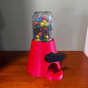 Puede incluir: Un dispensador de caramelos rosa con un frasco de vidrio transparente lleno de caramelos de colores. El dispensador tiene una manija negra y una pequeña abertura para dispensar caramelos.