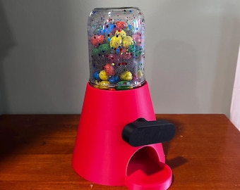 Op de afbeelding: Een roze snoepdispenser met een glazen pot gevuld met kleurrijke snoepjes. De dispenser heeft een zwarte hendel en een kleine opening om snoepjes uit te geven.