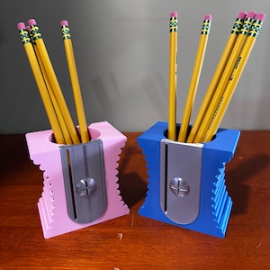 Könnte beinhalten: Zwei Bleistifthalter mit integrierten Bleistiftspitzer. Einer ist rosa und der andere blau. Jeder Halter hat sechs gelbe Bleistifte mit grünen und rosa Radiergummis.