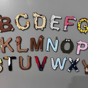 May include: A set of colorful alphabet magnets featuring animal illustrations. Each letter is decorated with a different animal, such as a giraffe, a zebra, a narwhal, and a penguin.