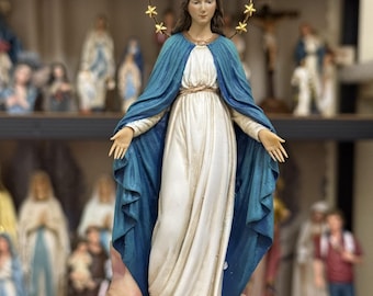 Peut inclure: Statuette religieuse d'une femme en robe blanche et cape bleue, avec une auréole d'étoiles dorées. La figurine est tenue devant une étagère avec d'autres statues religieuses. La base est un globe bleu avec un cœur rouge.