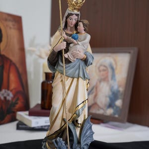 May include: A religious figurine depicting a figure wearing a gold crown and cream-colored robes, holding a child who is grasping a blue orb. The figure holds a tall gold cross staff and stands atop a gray dragon-like creature on a base.