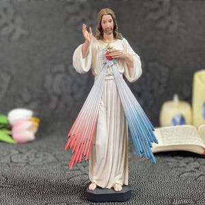 Può includere: Una statua religiosa di Gesù Cristo, vestito con una veste bianca, con luce rossa e blu che emana dal suo petto. La statua ha un'espressione serena e si trova su una base nera. Un cuore è visibile sul suo petto.