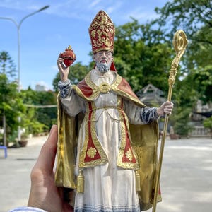 May include: A detailed statue of a religious figure, wearing a red and gold mitre and cape over a white robe. The figure holds a golden staff and a red heart-shaped object. The statue is outdoors with a blue sky and greenery in the background.