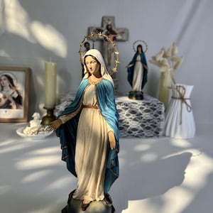 May include: A religious figurine of the Virgin Mary standing on a globe, wearing a cream-colored gown and a blue mantle. A gold-toned halo with star accents encircles the head. The figure is positioned on a dark, circular base with a red accent.