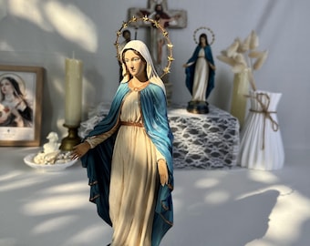 May include: A religious figurine of the Virgin Mary standing on a globe, wearing a cream-colored gown and a blue mantle. A gold-toned halo with star accents encircles the head. The figure is positioned on a dark, circular base with a red accent.