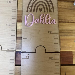 Può includere: Grafico di crescita in legno con segni di altezza incisi e il nome "Dahlia" in rosa. Presenta un design ad arco laserato e un ritaglio per la misurazione. Le misure sono in centimetri.