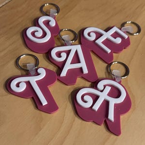 Peut inclure: Cinq porte-clés roses avec des lettres blanches en cursive formant le mot "STAR". Chaque porte-clés a un anneau argenté et une boucle en plastique transparente.